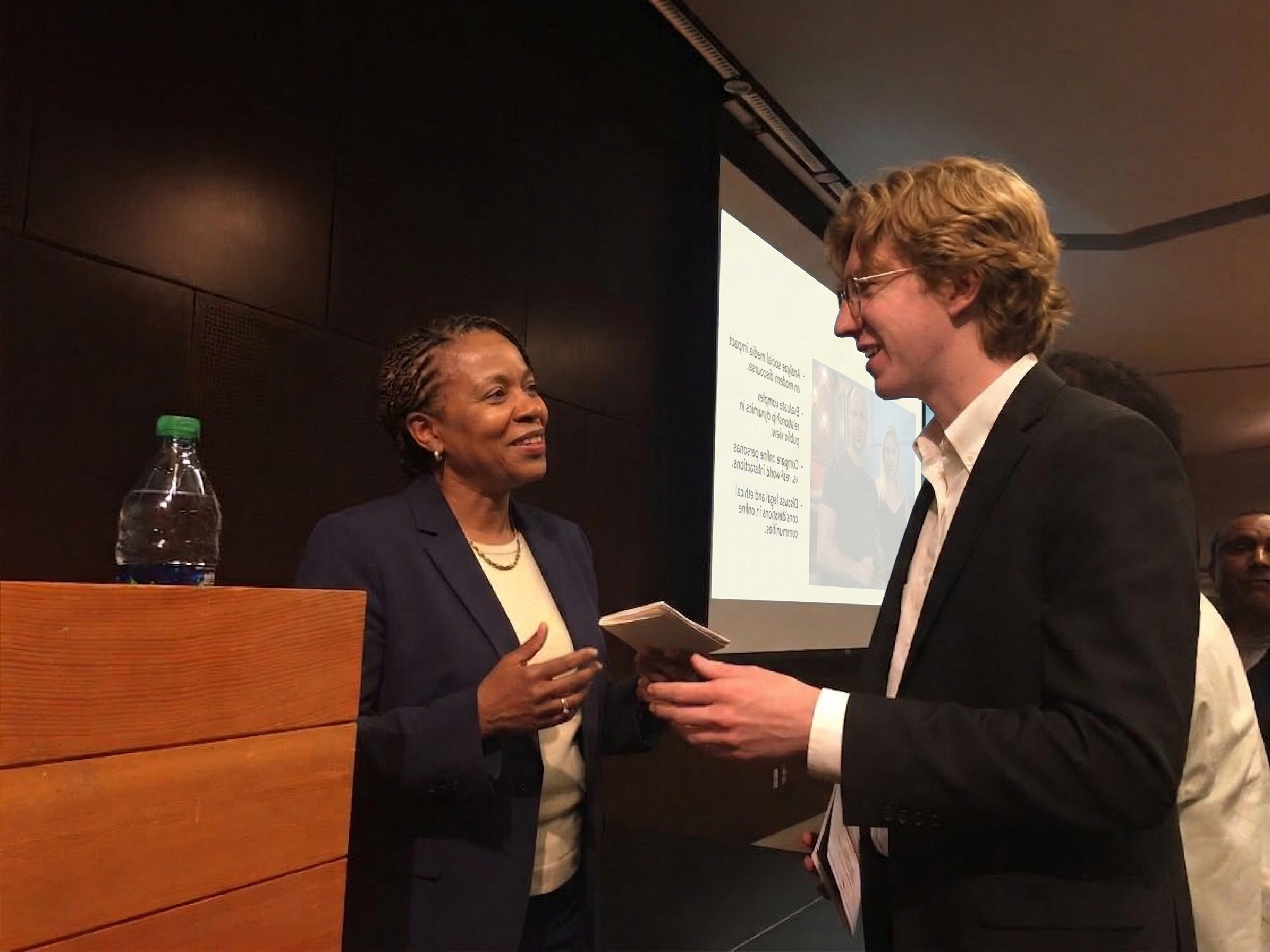 A speaker and attendee talking at a lecture podium, with a projection screen in the background
